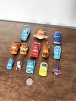 Photo showing 13 Pixar Cars toy vehicles on a wooden surface, including Lightning McQueen, Mater, and an orange airplane. Coin placed for scale.