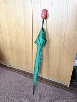 Full view of the closed umbrella showing the green fabric and tulip-shaped handle leaning against a wooden surface.