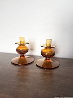 Pair of amber glass bulbous candle holders shown side by side on a wooden surface against a white backdrop.