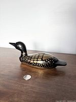 Side view of brass loon piggy bank with detailed feather pattern, displayed on wooden surface with quarter for scale.