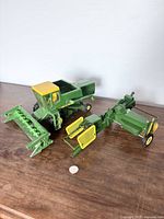 Photo shows a green and yellow diecast combine harvester with front header attachment and another farming implement, placed on a wooden surface with a coin for scale.