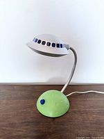 Front view of the vintage plastic UFO space lamp showing the dome-shaped white lamp head with blue windows, green textured base, and flexible neck.
