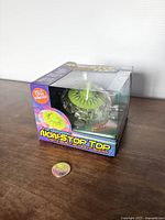 View of the boxed spinning top toy showing the clear plastic window, green top with light holes, and coin for scale.