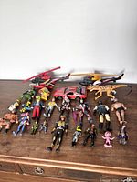 Wide shot of the entire lot showing two helicopters, a red toy car, dinosaur figure, and multiple action figures on a wooden surface.