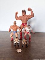 Five vintage WWE wrestling action figures displayed upright on wood surface with a coin for scale.