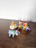 Set of 4 holiday Winnie the Pooh themed ornaments displayed on wooden surface: Pooh with gift, Pooh with Eeyore, Eeyore alone, and snow globe with Pooh and friends.