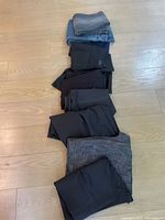 10 folded pairs of pants and jeans in various colors including black, navy, grey, and denim, stacked on a wooden floor
