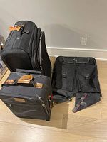 Three black fabric luggage pieces on floor including two upright suitcases with retractable handles and one garment bag with buckle-closed pockets.