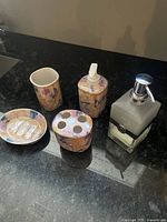 Ceramic bathroom accessories including soap dish, toothbrush holder, soap dispenser, cup, and a glass soap dispenser on black counter.