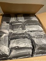 Large cardboard box filled with numerous black mesh cases individually wrapped in clear plastic packaging, stacked closely together.