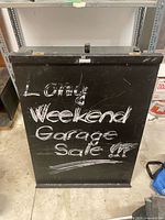 Front view of the black metal chalkboard sign with chalk writing advertising a long weekend garage sale.