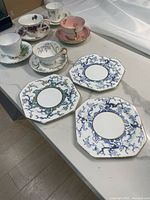 Five decorated vintage teacups on matching saucers arranged on a table, with three octagonal side plates showing blue and gold floral designs in front.