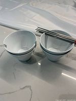 Four white porcelain food bowls stacked, each has a notch for chopsticks, accompanied by four metal chopsticks resting on bowls.