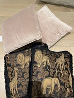 Four throw pillows arranged on floor, two beige velvet and two black with gold animal print.