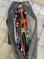 Photo of wooden croquet mallets, painted handles, balls and metal wickets inside black carrying bag on stone surface.