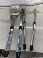 Photo of stainless steel spatula, fork, and tongs with black handles all wrapped in protective plastic.