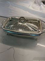 Closed view of ornate silver plated serving tray with embossed lid and handle knob over Pyrex glass dish.