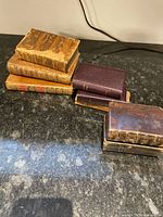 Overview of 7 of the leather bound miniature books stacked in two piles on a dark surface, showing wear and aging on the bindings.