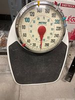 Front view of Metro mechanical bathroom scale showing large circular dial with red pointer and black rubber platform.