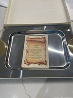 Rectangular stainless steel serving platter with attached gold-finished handles placed inside an open beige box. A parchment-style certificate of guarantee rests inside the platter.