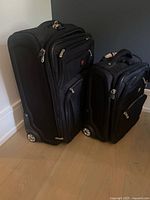 Photo showing two black Swiss Gear suitcases side by side on a hardwood floor; one carry-on size and one larger 25-inch piece, each with several zippered pockets and wheels.
