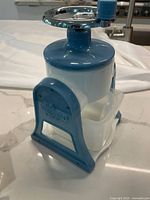 Blue and white plastic ice shaver unit viewed from side, showing hand crank and collection container with blue support frame.