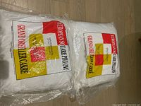 Two white European square pillow inserts in original clear plastic packaging with red, yellow, and black labeling, showing brand and dimensions.