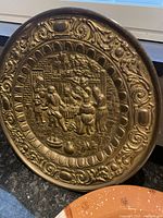 Frontal image showing the entire embossed brass wall plate with detailed village or tavern scene and ornate border patterns.
