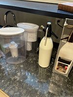 Three items on counter: two clear plastic Brita water dispensers with white lids and an off-white soda siphon with metal parts next to an original cartridge box.