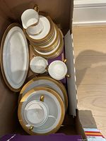 Photo showing stacked plates, cups, and serving platter with white and gold rims in a storage box.