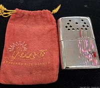 Front view of JON-E silver tone hand warmer alongside red flannel pouch with gold text and sunburst logo.