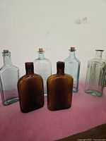 Six antique medicine bottles arranged on pink fabric. Four blue-tinted glass bottles with embossed writing, two brown molded glass bottles, all with cork stoppers.