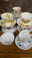 Full view of 8 assorted fine bone china teacups and saucers stacked and arranged with 1 sugar bowl