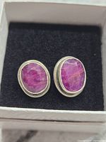 Pair of oval ruby stud earrings set in sterling silver displayed in black velvet box under normal light. Shows natural ruby inclusions and silver bezel setting.