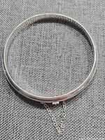 Full view of the silver hinged bangle on a fabric background, showing circular shape and attached safety chain.