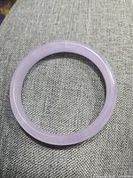 Photo showing the light purple amethyst jade bangle bracelet laid on a gray textured surface, highlighting the smooth polished finish and visible hairline crack.
