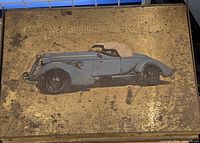 Top lid of the antique metal box showing a faded image of a vintage convertible automobile against an aged metal background.