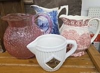 Four assorted pitcher jugs placed on a wooden surface: large pink textured glass pitcher, vintage blue and white scenic ceramic pitcher, vintage pink and white floral patterned ceramic pitcher, small white ceramic milk pitcher with diamond MILK label.