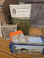 Assorted vintage tins and small boxes stacked on a wooden surface including Laura Secord Candies tin, Rosemary Tea tin with floral decoration, cardboard boxes, and a glass medicine bottle.