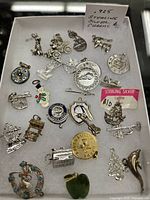 Photo displaying the full assortment of sterling silver charms laid on a white background with a labeled sign noting '.925 Sterling Silver Charms 4/1'. Charms include varied designs: carriage, musical instruments, holiday symbols, and state of Kentucky among others.