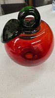Front angle photo of the red blown glass apple with green leaf and looped stem on white table surface.