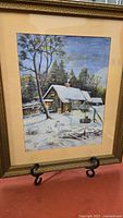 Front view of framed winter landscape painting depicting snow-covered house and trees, placed on display stand.