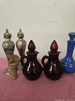 Photo showing assorted decorative bottles including two ornate silver-tone bottles, two maroon glass bottles with red stoppers, and one cobalt blue bottle.