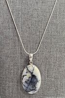 Front view of necklace with oval dendritic opal pendant on silver snake chain against grey fabric background.
