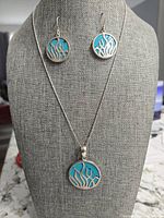 Photo of the pendant and matching earrings displayed on a gray fabric bust. Shows the turquoise blue acrylic with silver seagrass overlay design. Earrings have hook backs, pendant on chain.