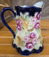 Left side angled view of the porcelain pitcher showing pink roses and gold scroll details with blue handle and rim.