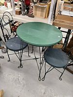 Photo showing entire bistro set including round green table with metal legs and two black metal chairs with circular metal seats and decorative backrests. Items arranged upright showing shape and condition.