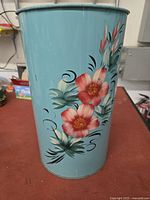 Front and side view of the tall teal tin garbage can showing hand-painted pink and red floral design with green leaves.