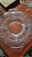 Top view of the round glass serving plate showing the intricate silver etched floral and scrollwork design with one handle visible.