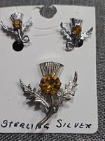 Front view of sterling silver Scottish thistle brooch and matching screw-on earrings on white card. Amber-colored rhinestones in center. Detail of metal leaf design.